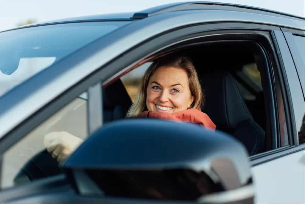Kvinne i førersetet smiler ut av det åpne vinduet.