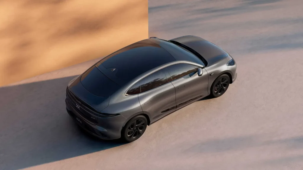 Sølvfarget elbil med glasstak parkert ved lys vegg.