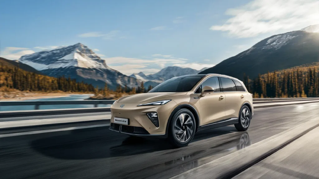 Beige elbil kjører på fjellvei ved innsjø, snødekte topper bak.