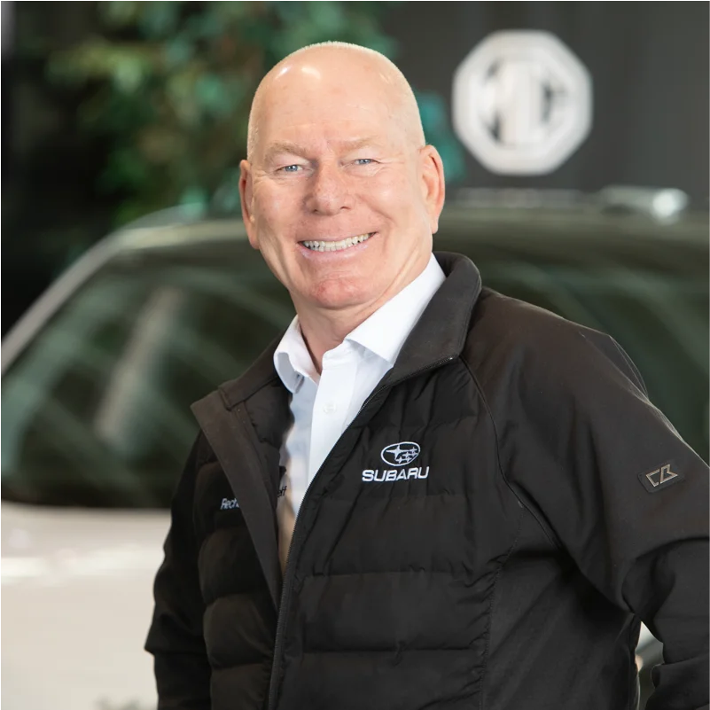 Smilende mann i svart jakke foran hvit bil innendørs.