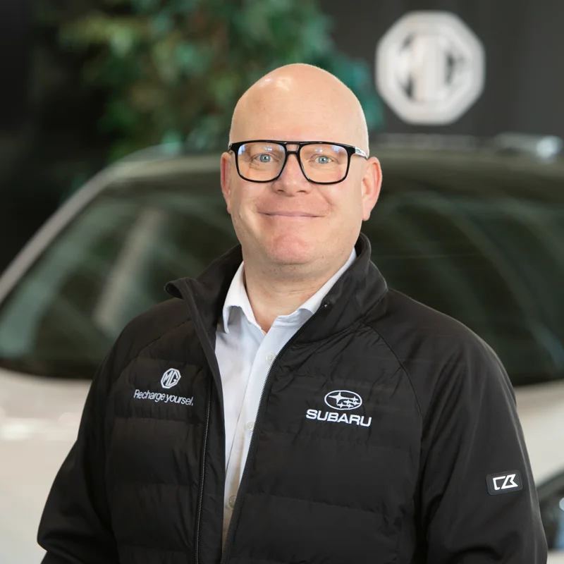 Mann med briller foran hvit bil inne hos bilforhandler.