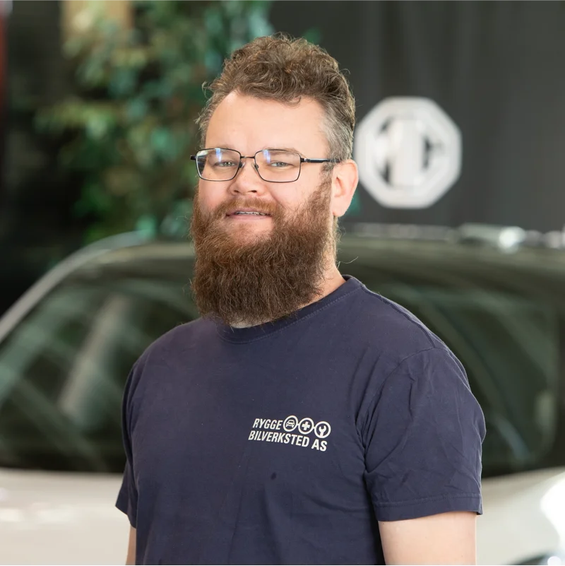 Mann med briller og skjegg foran uklar bil og busker.