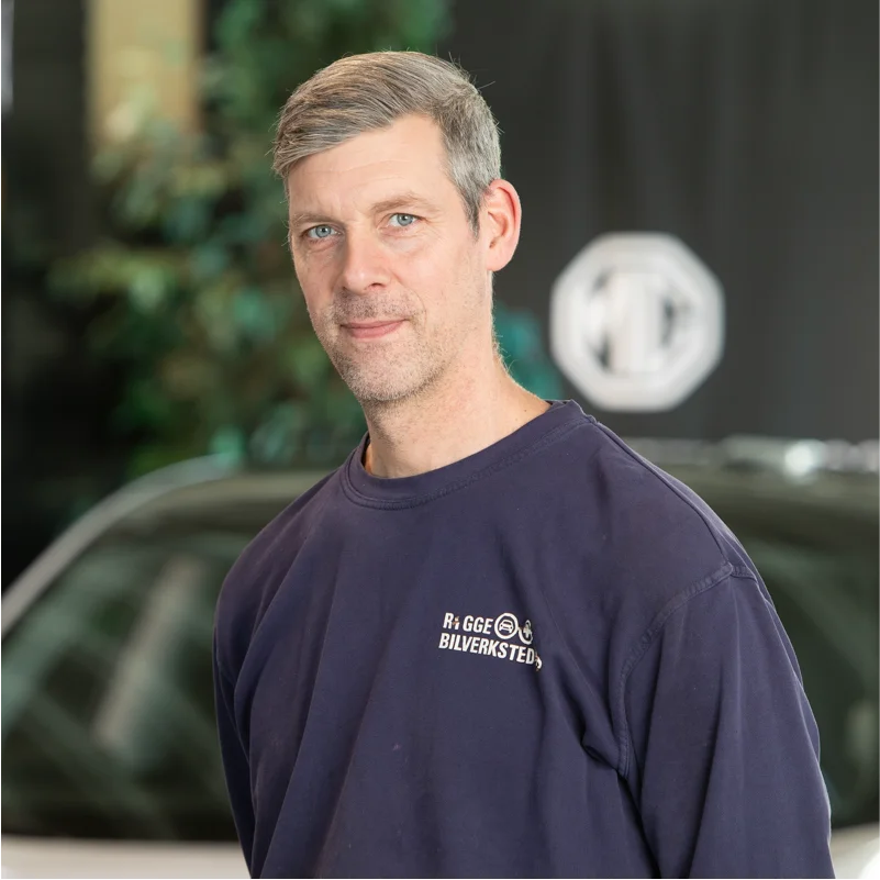 Mann med grått hår foran bil og logo, innendørs.