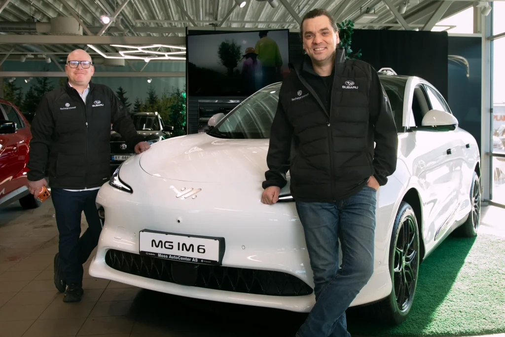 To menn i svarte vester ved hvit bil i et showroom.